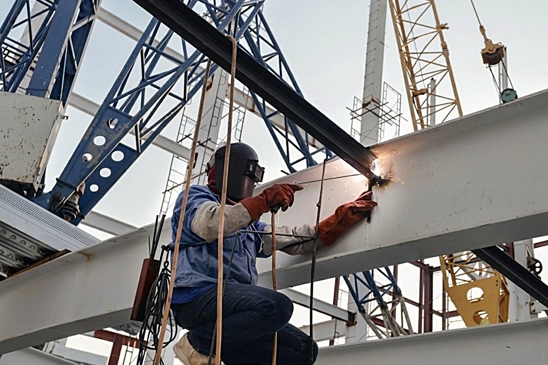 Welder working on a structural steel beam at height with a crane in the background